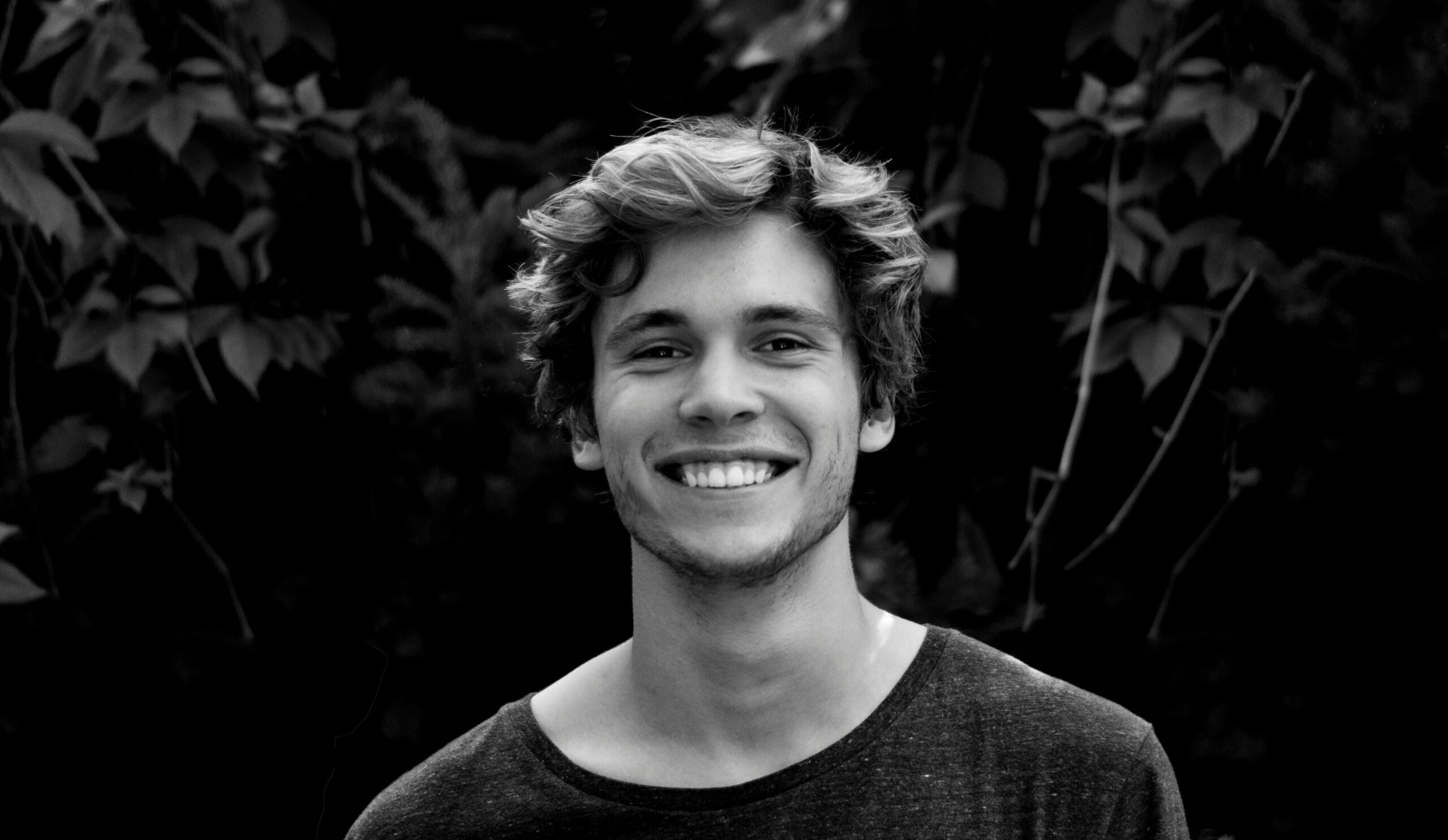 A black and white portrait of a young man smiling outdoors with natural background.